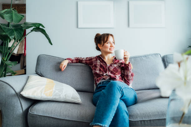 istock Young smiling woman sitting on sofa and looking side up while drinking coffee or tea. Young brunette woman relaxing after housekeeping, home cleaning. Portrait of relaxed female resting at home 1558511200