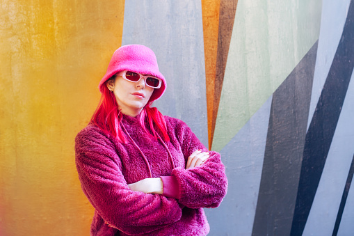 Urban autumn winter street fashion. Kawaii vibes. Young woman with pink hair and sunglasses in a magenta fluffy sweatshirt and bucket hat crossed arms on a graffiti wall background. Hipster lifestyle