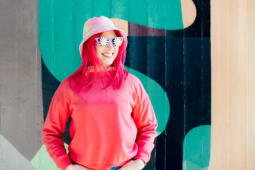 Happy smiling woman in sunglasses with flowers stickers. Woman with pink hair,bucket hat and magenta sweatshirt posing on graffiti wall background. Vanilla Girl. Kawaii vibes. Candy colors design.