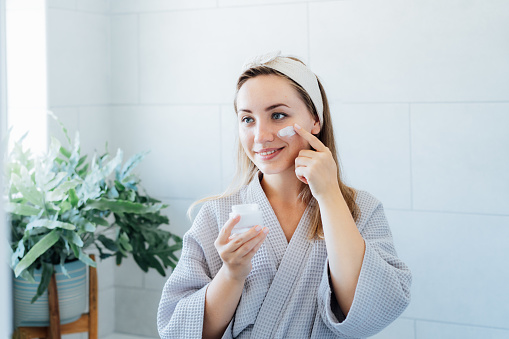 Smiling young woman with skin problems in bathrobe takes care face skin at home. She looking in the mirror and aplying cream on the face in bathroom. Acne skin care every day treatment process