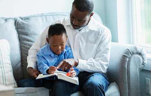 A young multiracial boy sitting with his father at home on a couch, reading a book. Father is a mature African-American man, in his 40s. His son is mixed race Asian and African-American, 5 years old.