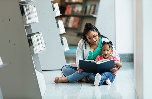 Mixed race little girl reading with mother in library