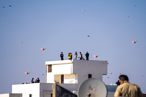 Jaipur, Rajasthan - 14th Jan 2023: young men women children standing on roof of concrete house flying kites fighting on sankranti uttarayan independence day against clear blue sky in India