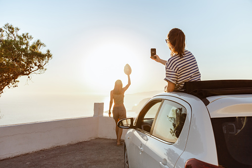 Rear view of a woman photographing her friend from the open roof of her car