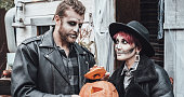 Scary love family couple man, woman celebrating halloween with pumpkin jack-o-lantern. Terrifying black skull half-face makeup,witch costumes,stylish images,jacket,hat.Horror,fun at photoshoot, holiday party near barn on street