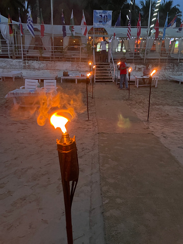 Stock photo showing twilight view of flaming torches lining a carpet on a sandy beach leading to staircase entrance of beach restaurant with terrace lined with international flag.