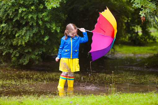 Little girl playing in rainy summer park. Child with colorful rainbow umbrella, blue coat jumping in muddy puddle, walking in the rain. Kid having fun in autumn shower. Outdoor activity by any weather