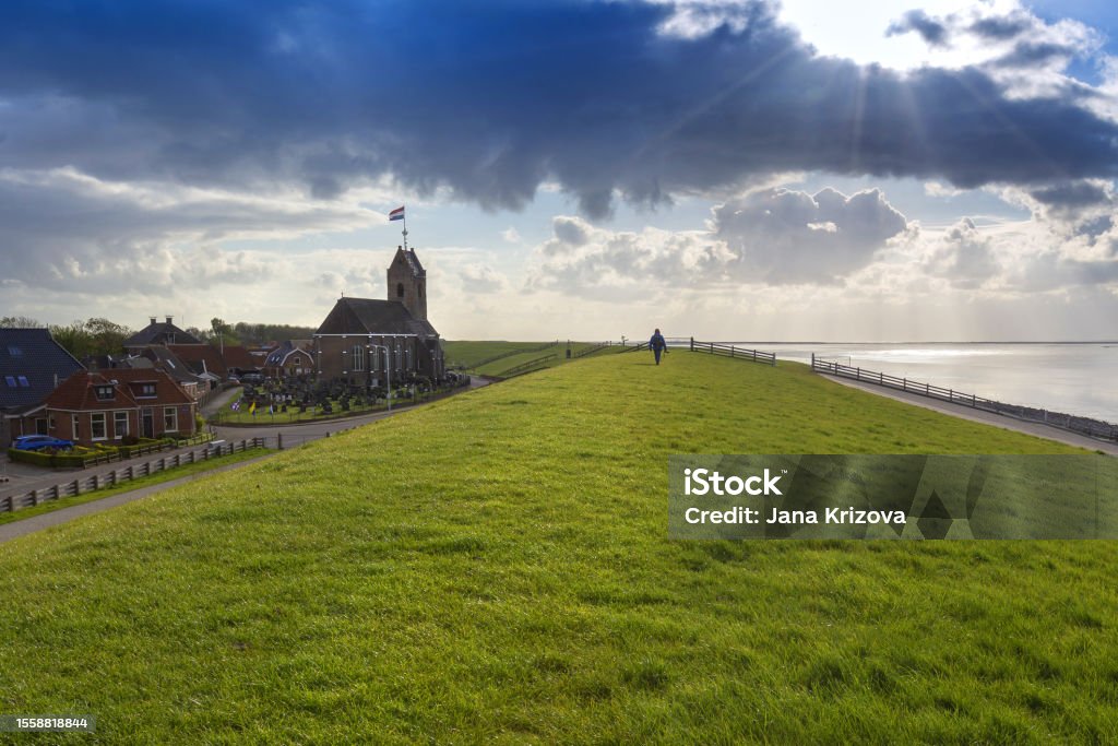 Holland - Hindeloopen - coast, high coastal wall overgrown with grass. On the right side of the sea and on the left side houses Hindeloopen and a wooden church. Dramatic sky with clouds. Netherlands Stock Photo