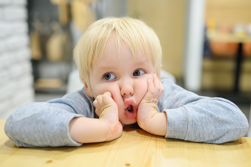 Funny emotional child is at a table in a cafe. Cute little boy sitting in restaurant and waiting for his order. Kid tired of waiting and is making faces. Sincere behavior of children in public places