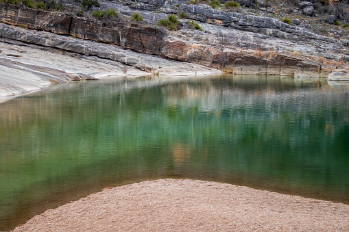 Geological granite stone and limestone wall mirrored in a green pond shoreline in Pedernales Falls State Park as part of the Texas Hill Country