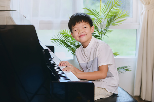 Cute happy smiling little Asian kid boy playing piano in living room at home, Elementary school child having fun with learning to play music instrument, Music education concept