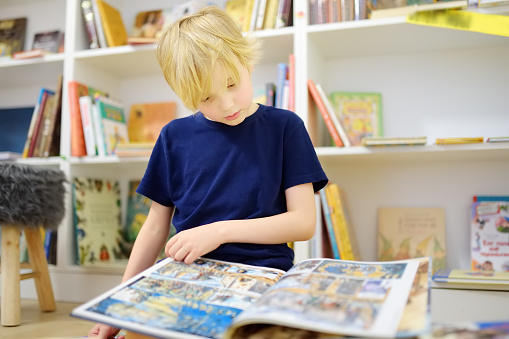 A preteen boy leafing through a book while sitting at the bookshelves at home, in a school library or bookstore. Smart kid reading comics or adventure book