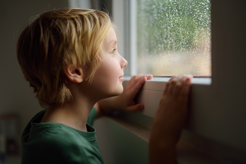 Portrait of preteen boy standing at the window. Child watching the rain outside. Faith and hope concept.
