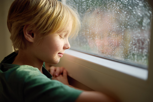 Portrait of a cute preteen boy standing in meditation or prayer at the window. Teaching emotional control to teens. Expression of feelings in healthy way. Development of emotional intelligence.