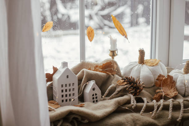 Cozy Autumn. Stylish pumpkin pillows, fall leaves, candle, lights and cute buildings decoration on brown scarf on windowsill. Autumn hygge, fall home decor. Happy Thanksgiving stock photo