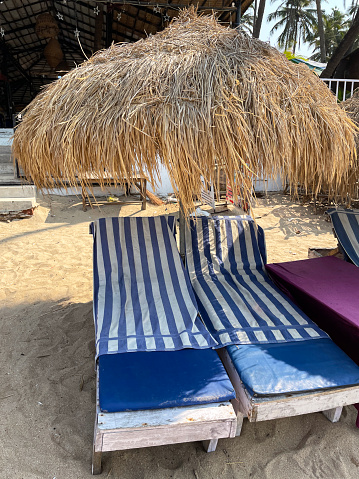 Stock photo showing close-up view of row of blue cushioned, wood sun lounger beds covered with blue and white striped towels under thatched parasol sun umbrellas on beach holiday at Palolem Beach, Goa, India.
