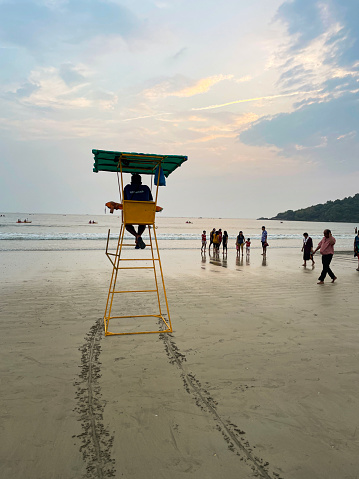 Palolem Beach, Goa, India - November, 27 2022: Stock photo of a lifeguard watching the Arabian Sea from an observation chair on Palolem Beach, Goa, India with view of bathers swimming and boats floating on the waves.