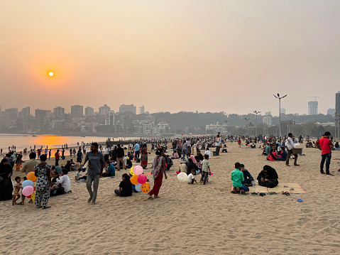 Girgaon Chowpatty, Mumbai, India - March, 5 2023: Stock photo showing close-up view of crowd of tourists, holidaymakers and locals on beach and in the sea to enjoy the sunset. Mumbai is an extremely overcrowded city with few public open spaces so the beach has become a popular location to meet with friends and family.