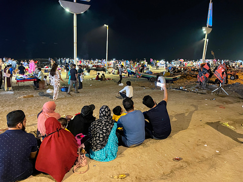 Girgaon Chowpatty, Mumbai, India - March, 4 2023: Stock photo showing close-up view of crowd of tourists, holidaymakers and locals on the beach at night with illuminated gift shop kiosks. Mumbai is an extremely overcrowded city with few public open spaces so the beach has become a popular location to meet with friends and family.
