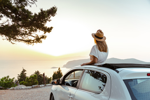 Cheerful young woman in an open-top car. She is enjoying the sunset and looking at the sea