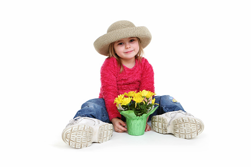 Front view of little girl in jeans and pink shirt  with straw  hat and adult oversized tennis shoes.  She's sitting down holding flower pot with plant in front of her. Full body showing on white background. Part of an on-going series.