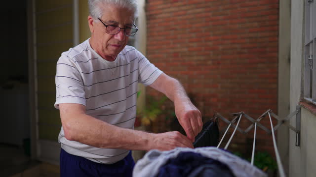 Senior man putting clothes to dry on hanger in home backyard. Independent elderly person doing domestic weekly routine at casual humble residence