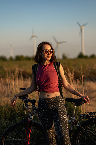 Female portrait of beautiful woman with mountain bike outdoors in the field during sunset. Lifestyle photo with beautiful people, person wear sunglasses. Photography with turbines of wind farm, green energy technology