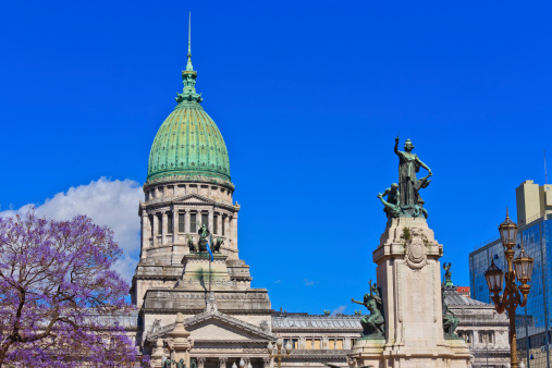 The monument of the two congresses (Monumento a los dos congresos) with the Congress Palace (national parliament) or Palacio del Congreso de la nacion argentina in downtown Buenos Aires in the background and blue sky above; blooming jacaranda tree covering part of the congress palace