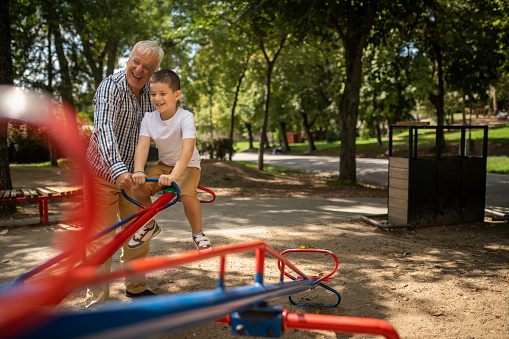 Little boy playing on seesaw with his grandfather in public park playground.
