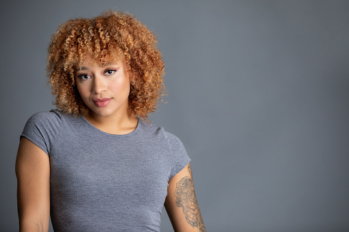 Portrait of edgy fashion model with piercings and tattoos shot on gray studio background. Model is mixed race young woman in her twenties with natural curly hair dyed strawberry blonde.