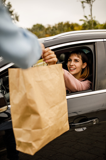 Cut out shot of unrecognizable service person handing a paper shopping bag to a customer in her car at the curbside pickup.