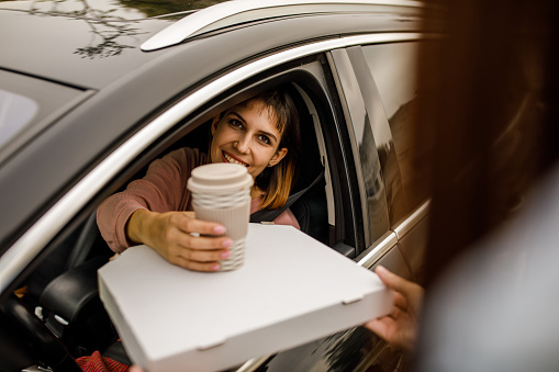 Selective focuc shot of young woman sitting in her car, in driver's seat, smiling at service person who is handing her a coffee and takeaway pizza at the drive through.