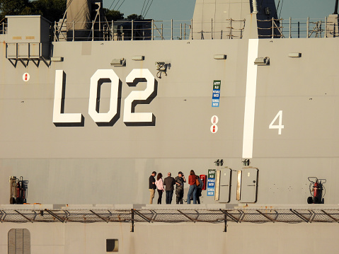 A high-ranking naval officer with people in civilian clothes on the deck of HMAS Canberra, an amphibious assault ship of the Royal Australian Navy.  A US Navy ship and two Republic of Korea Navy ships are also docked in preparation for the commissioning ceremony of USS Canberra on 22 July 2023.  This image was taken from Mrs Macquarie's Chair shortly before sunset on a cold, sunny day on 20 July 2023.
