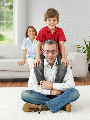 Boy sitting on his father’s shoulder and smiling to the camera in a modern house