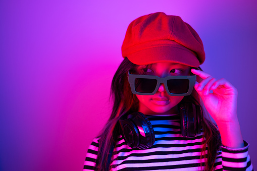 Portrait of an Asian girl wearing green sunglasses with headphones around her neck and a red hat with blue and red neon lights.