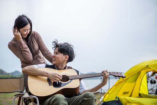 Young Asian couple play guitar and sing happily in nature on weekend. Men and women go on a camping trip. vacation Taking care of each other's love for each other.