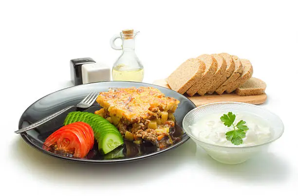 Traditional Bulgarian dish arranged in a black ceramic plate with fresh sliced tomato and cucumber with a sprig of parsley, slices of bread, bowl of yogurt, a bottle with olive oil and containers for salt and pepper around isolated on a white background