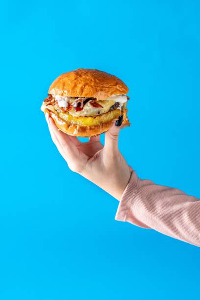 istock Female hand grasping a freshly-made chicken burger on a blue background. 1559917705