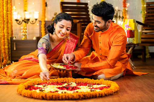Happy indian husband helping to wife decorating rangoli with flowers for Diwali festival celebration while sitting on floor at home - concept of relationship, bonding, and religious ceremony