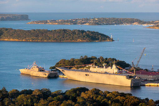 USS Canberra (LCS 30) and behind her, HMAS Canberra (L02), docked at Garden Island in Sydney Harbour.  USS Canberra is an Independence Class combat ship of the US Navy.  She is in port for a commissioning ceremony on 22 July 2023.  She is adorned with red, white and blue bunting and maritime flags.  HMAS Canberra is an amphibious assault ship of the Royal Australian Navy.  She is adorned with maritime flags.  In the distance is the Sydney Heads and the Pacific Ocean. The areas of eucalyptus trees form parts of the Sydney Harbour National Park.  This image was taken from the 29th floor of Chifley Tower on a cold, sunny afternoon at sunset on 21 July 2023.