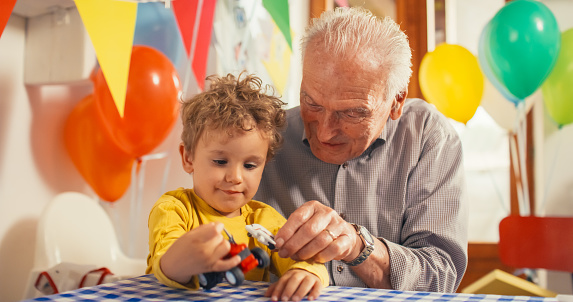 Heartwarming Moment Shared Between a Senior Man and his Grandson: Happy Toddler and his Grandpa Playing with Truck and Plane Toys Together in a Kitchen Decorated with Balloons for a Birthday Party