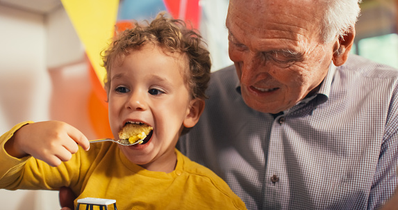 Portrait of a Little Cute kid Sitting on His Grandfather's Lap While Eating a Delicious Birthday Cake. Male Toddler Enjoying the Sweet Snack After Playing for Long at a Party