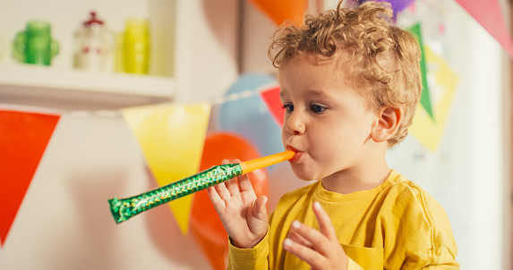 Colorful Happy Atmosphere:  Portrait of Little Cute Boy Blowing a Party Whistle on his Birthday. Blond Male Toddler Focused on the Sound of a Party Blower. Concept of Innocence, Fun and Childhood