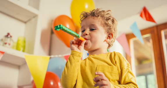 Low Angle Portrait of a Male Kid Using a Party Blower in a Living Room Decorated by Balloons. Little Cute Boy Blowing a Party Horn and Laughing on his Birthday. Happy Toddler Celebrating and Smiling