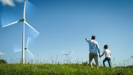 Father And Daughter Walking Through Modern Wind Farm. Professional Male Engineer Showing Next Generation Sustainable Green Energy Resources. VFX Graphics Edit Visualizing Wind Moving the Blades.