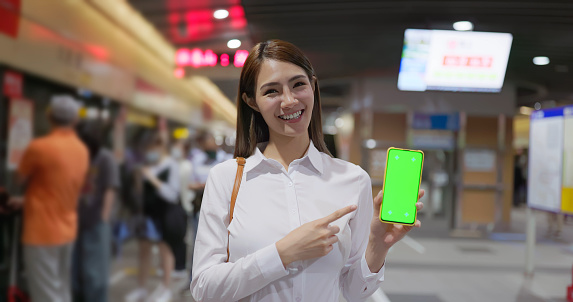 portrait asian business woman is showing green screen mobile phone on metro subway station platform
