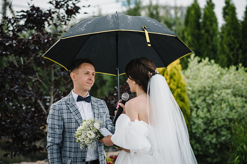 Bride says promise in microphone. Happy bride and groom exchanging wedding vows on ceremony in backyard banquet area. Newlyweds standing with bouquet in rain with umbrella. Fidelity in love. Back view
