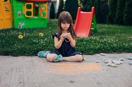 A child sits on the ground in the yard and draws with chalk. The girl chooses the next color for her art.