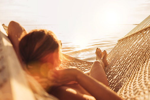 Young woman relaxing in wicker hammock on the sandy beach on Mauritius coast and enjoying sunset light over Indian ocean waves. Exotic countries vacation and mental health concept image.