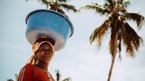 Palma, Mozambique – November 09, 2022: An African female holding a bucket on her head against palm trees in Palma, Mozambique